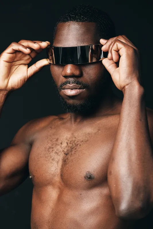 Black man with dark skin and muscular body wearing futuristic visor in creative conceptual visual style. Portrait of confident male model with bare chest and modern accessory on black background.