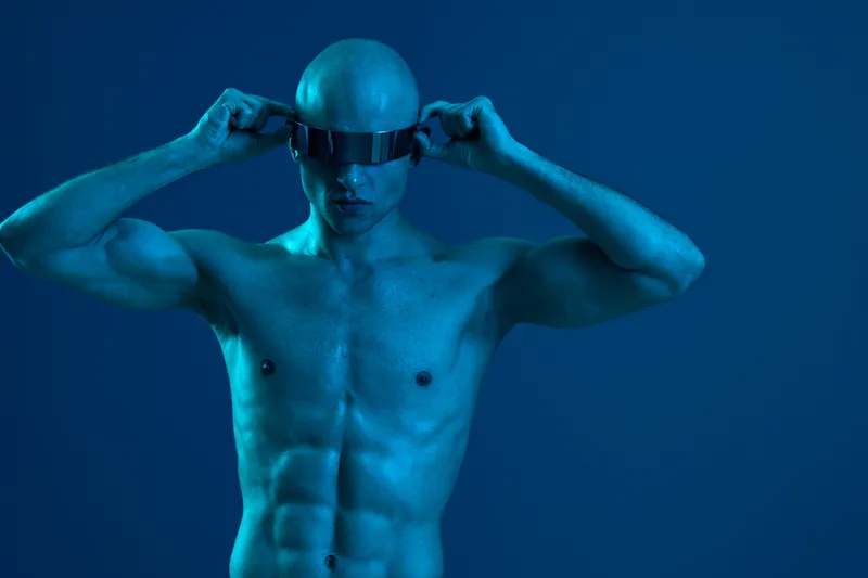 Athletic man wearing futuristic glasses poses confidently against a cool blue background, showcasing a striking black and white aesthetic in this captivating image.