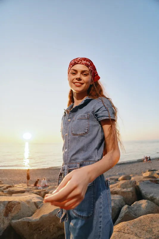 lifestyle woman in modern boho-western denim street style with red bandana smiling on beach at sunset in warm film color tones