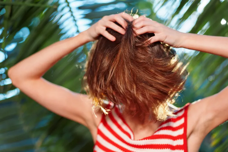 A person wearing a red and white striped sleeveless top adjusts their hair outdoors under bright sunlight, capturing a candid, carefree moment that conveys energy, youth, and playful style in a sunny