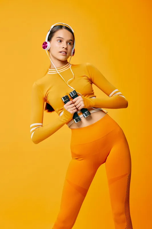Fitness woman in bright orange athletic wear holding dumbbells and wearing headphones, mid-workout against a vibrant orange background, showcasing energy, sport and focus.