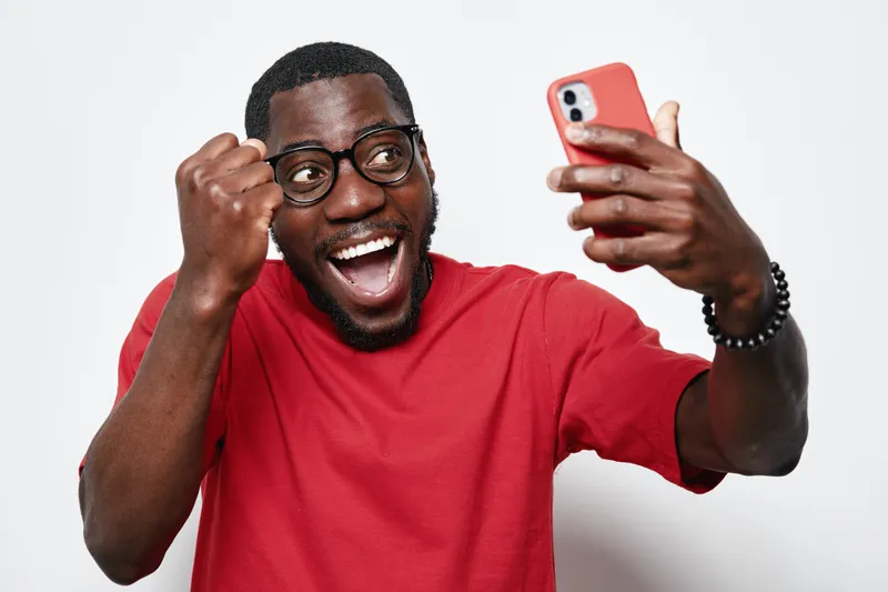 African American man with dark skin and glasses wearing red shirt taking selfie with smartphone, smiling and showing fist pump gesture on white background in studio.