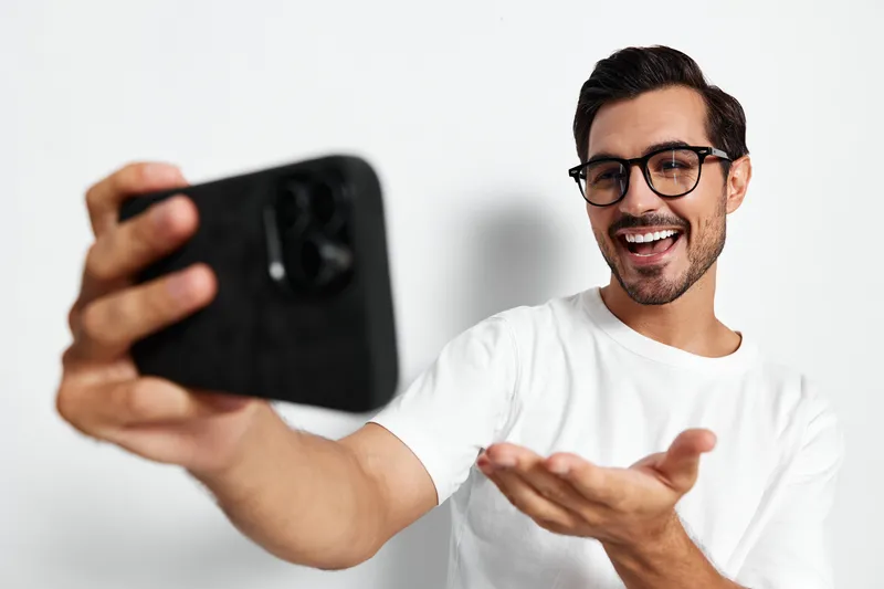 Cheerful smiling young man taking selfie with smartphone on plain solid background, wearing casual white t-shirt and glasses. Modern lifestyle concept.
