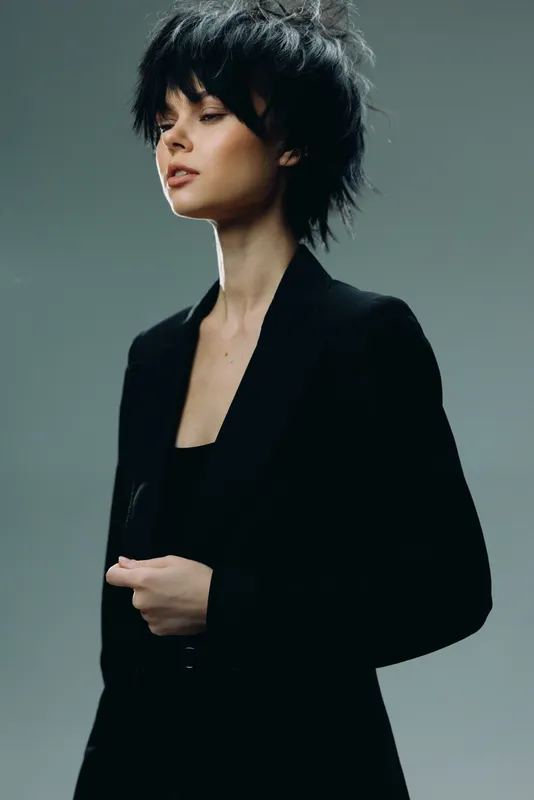 Stylish woman with short black hair wearing a black blazer, posing confidently against a gray background in a modern studio setting.