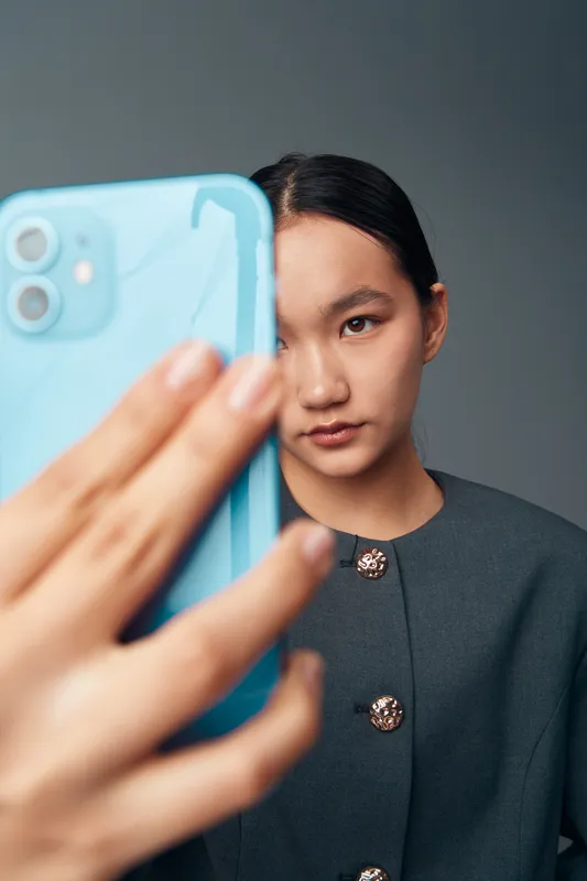 Young woman taking selfie with smartphone in blue case against gray background. Casual portrait with focused expression and dark hair wearing buttoned top indoors.