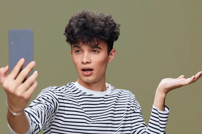 young man taking selfie with smartphone showing surprised expression wearing striped shirt on green background