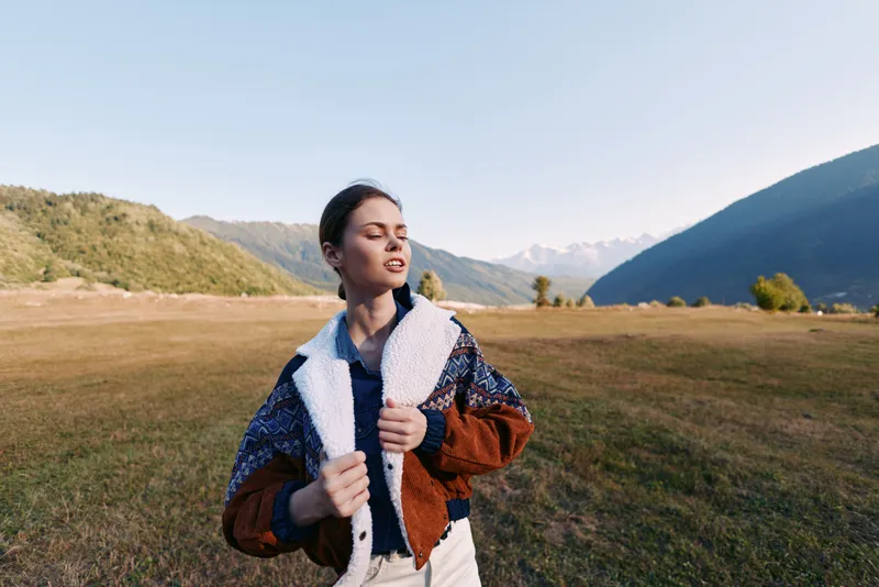 Woman portrait in nature standing on a wide meadow with mountains in the background, wearing a warm jacket during travel outdoors. Young person enjoying sunlight, scenic landscape and calm horizon.