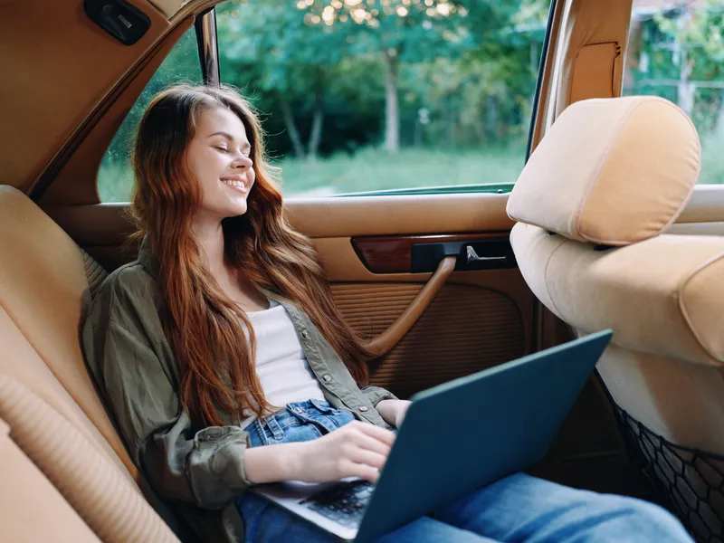 Young woman relaxing in a vintage car, working on a laptop with a serene smile, dressed casually in a green shirt and blue jeans, set in a serene green background.