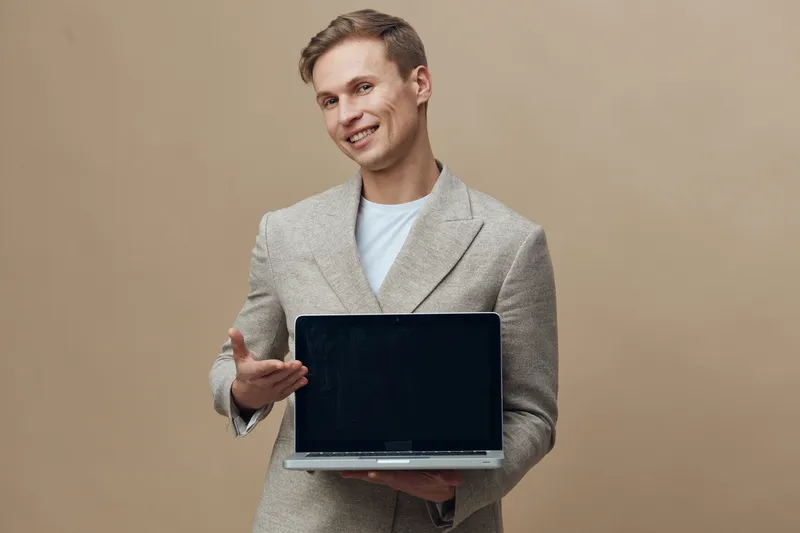 Smiling man holding open laptop computer in hand on plain beige background. Business casual attire, cheerful expression, modern technology. Concept: professional work, technology, communication, succe