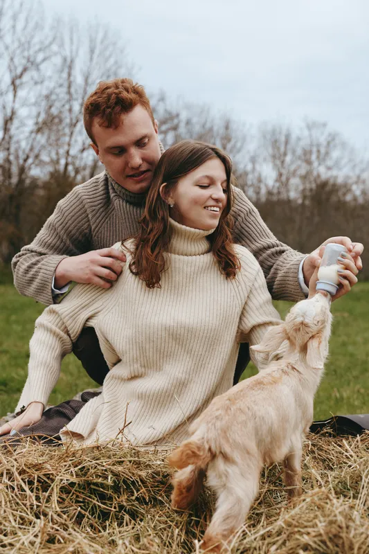 Young couple in cozy sweaters feeds a baby goat from a bottle while sitting on hay in a natural outdoor setting during autumn, enjoying a relaxed lifestyle moment together.