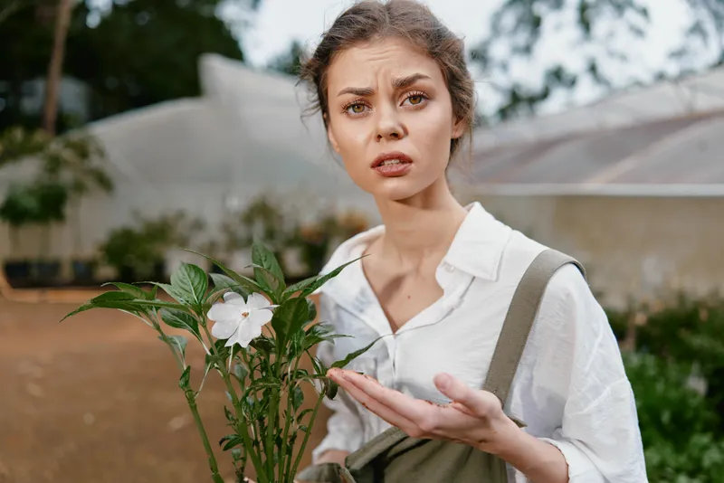 Young woman in garden expressing confusion while holding a plant, wearing casual attire, evoking emotions of curiosity and concern, surrounded by green foliage