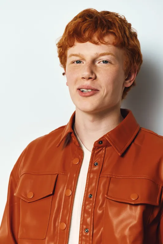 Young man with red hair wearing orange jacket and white shirt standing smiling against white background in studio portrait for casual fashion and lifestyle.