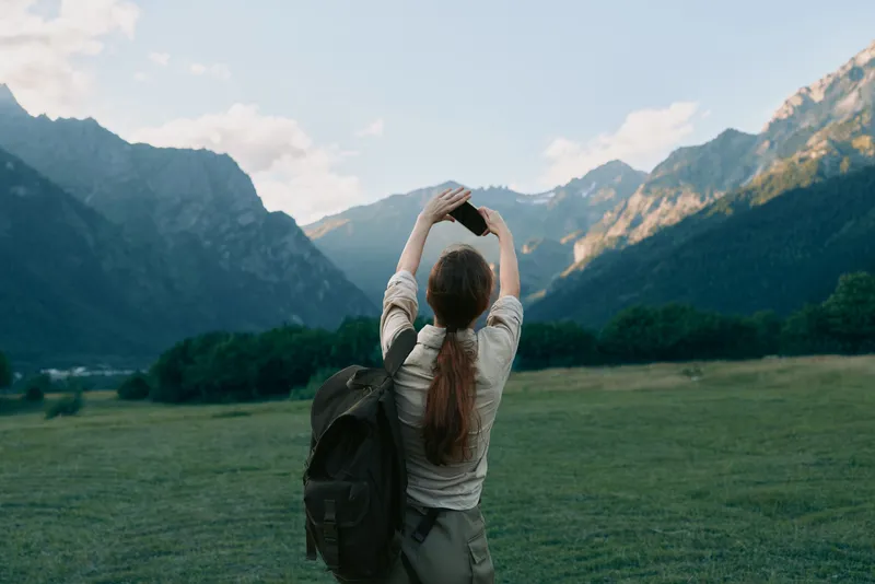 Woman traveler with backpack taking photo of mountain landscape, outdoor adventure, natural scenery, clear sky, green meadow, casual outfit, summer day