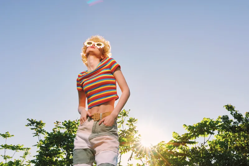 A woman stands outdoors under bright sunlight with a striped top and light shorts, captured in a retro film color palette. A vivid moment of carefree summer mood and casual style.