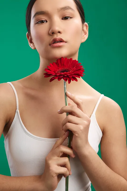 Asian woman holds a red flower near her face against a vivid green backdrop. The calm expression and minimalist styling create a polished, contemporary portrait with a fresh, positive mood.