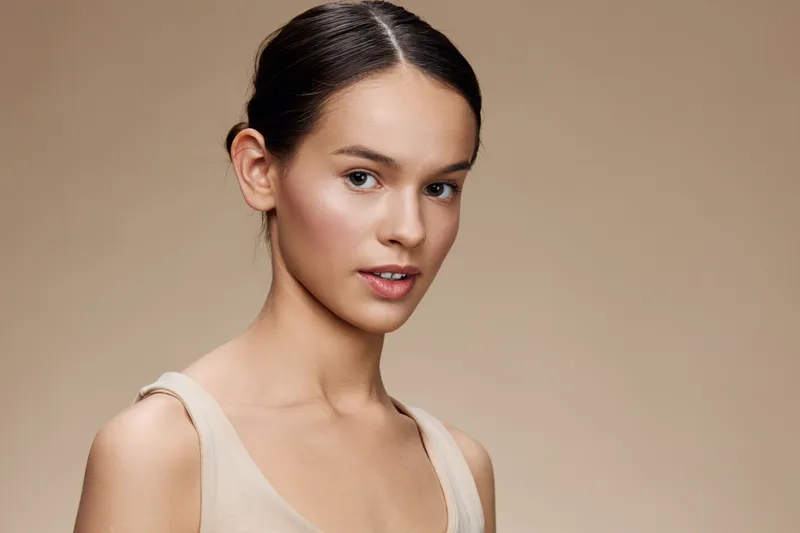 Natural beauty portrait of a young woman with light skin, wearing a simple beige top, showcasing a fresh, radiant look against a neutral background