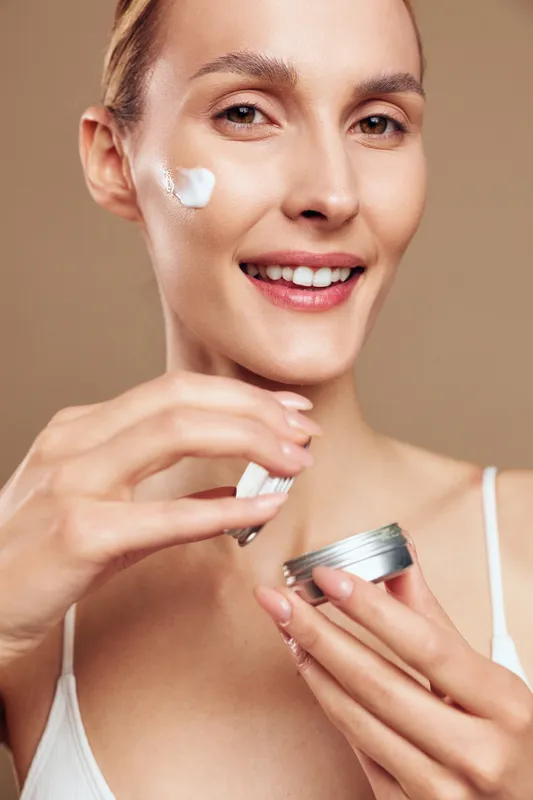 Smiling woman applying cream on her cheek, holding jar, natural skin care routine, close up portrait, soft lighting, beige background, healthy glowing complexion