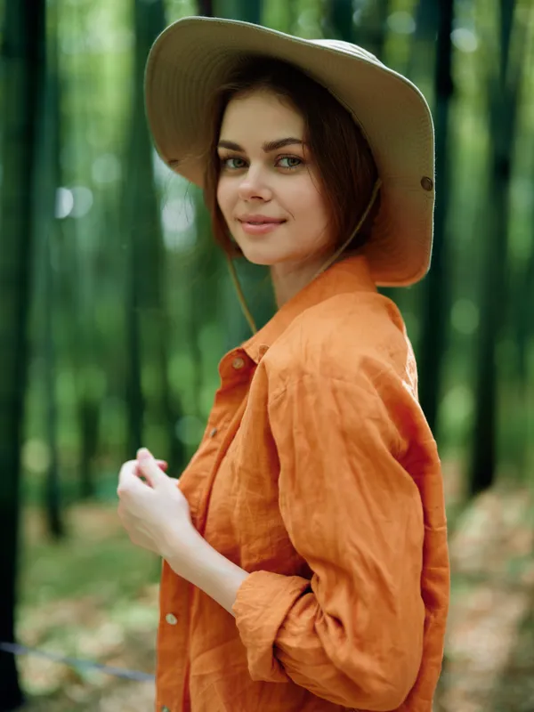 Woman portrait with hat in a green forest, orange linen shirt and gentle smile, outdoor nature scene with young female model in casual travel outfit and soft bokeh background.