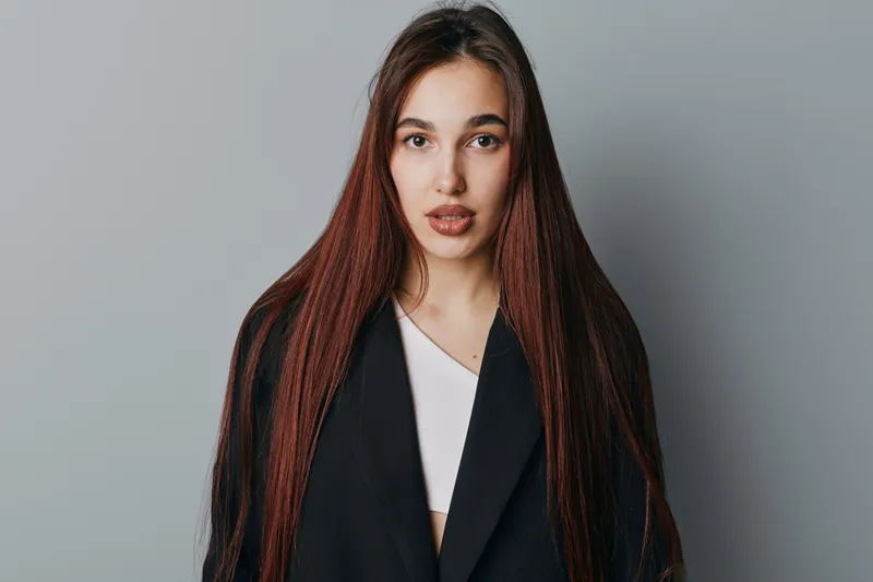Young woman with long straight hair wearing black coat and white top standing in front of gray background looking at camera with neutral expression in studio portrait.