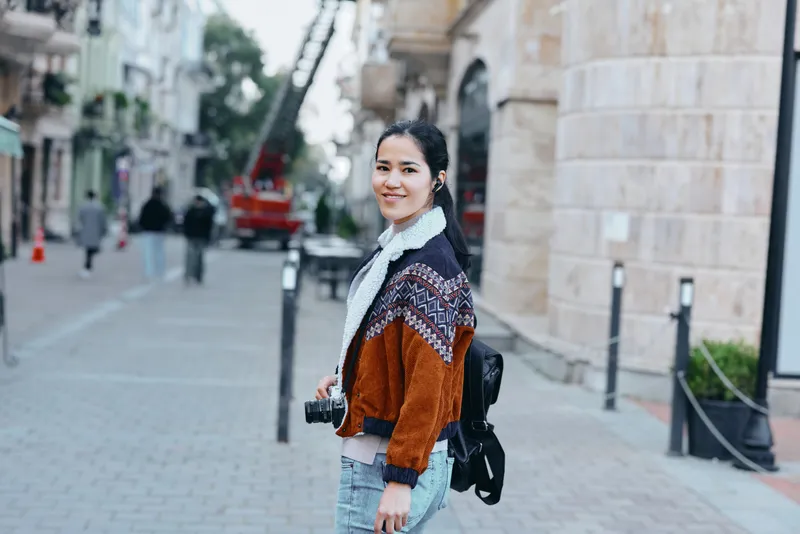 Woman with camera on a sunny city street, stylish casual fashion, denim jeans and backpack, smiling as she photographs the urban scene in a busy pedestrian corridor