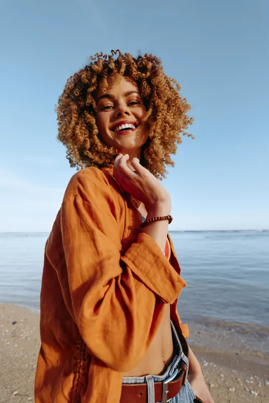 Smiling young woman with curly hair wearing an orange shirt stands on a sunny beach by the calm sea, enjoying a breezy moment and carefree mood.