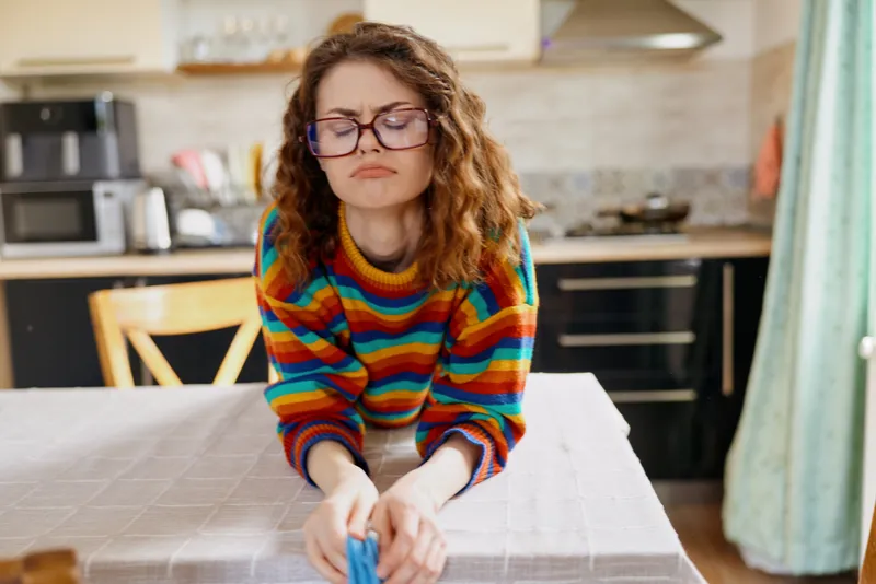 Woman in a colorful striped sweater leans over a kitchen table, hands arranging items, focused expression, indoor home setting, candid lifestyle shot, cozy domestic moment, casual morning routine
