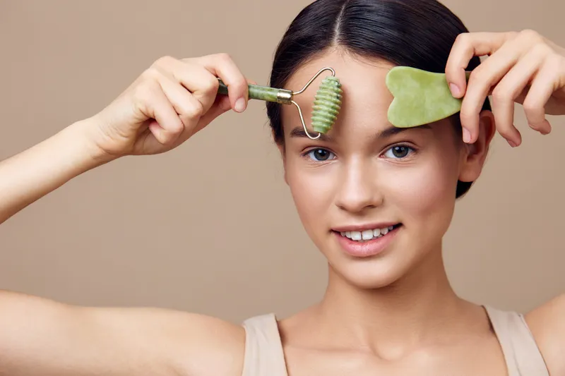 Young female model demonstrating a facial massager and gua sha tool, promoting skincare and relaxation, with a natural, serene expression against a soft beige background