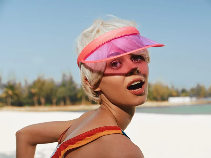 Vibrant portrait of a fashion beachgoer on a sunny day, wearing a pink visor and striped swimsuit, turning with a surprised expression against a clear blue sky.