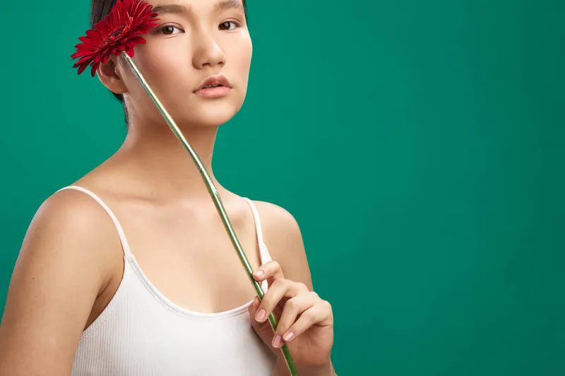 asian woman holds a red flower against a vibrant green background, creating a calm portrait that emphasizes elegance, simplicity, and contemporary style.