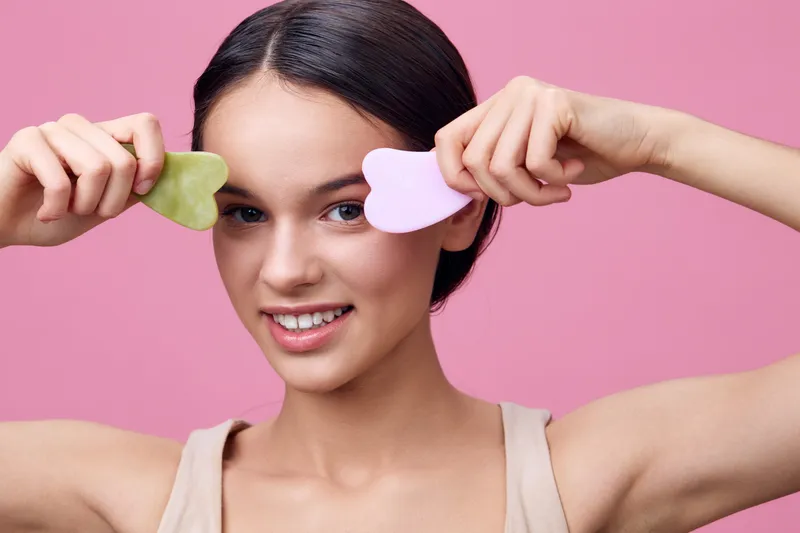 Young woman with natural beauty tools smiling, holding heart shaped gua sha stones in pastel colors against a soft pink background for a skincare routine