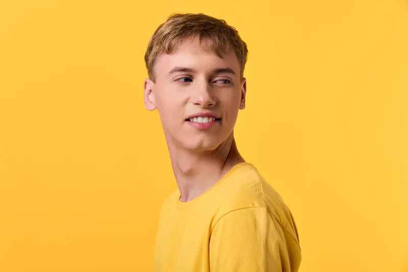 Young male in a bright yellow shirt smiles softly against a sunny backdrop, showcasing casual style, cheerful mood, and contemporary fashion vibe suitable for lifestyle or advertising shoots