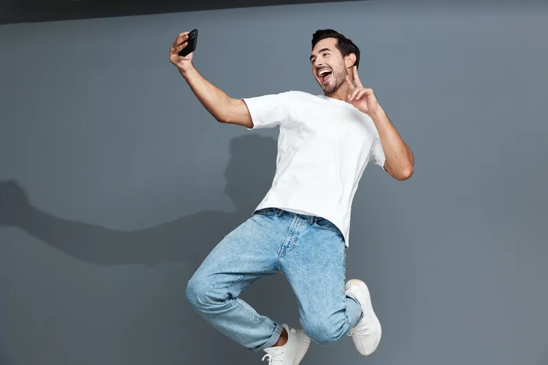 Full body cheerful happy young man taking selfie with smartphone, wearing casual white t-shirt and denim jeans, leaping against solid gray background. Lifestyle concept.