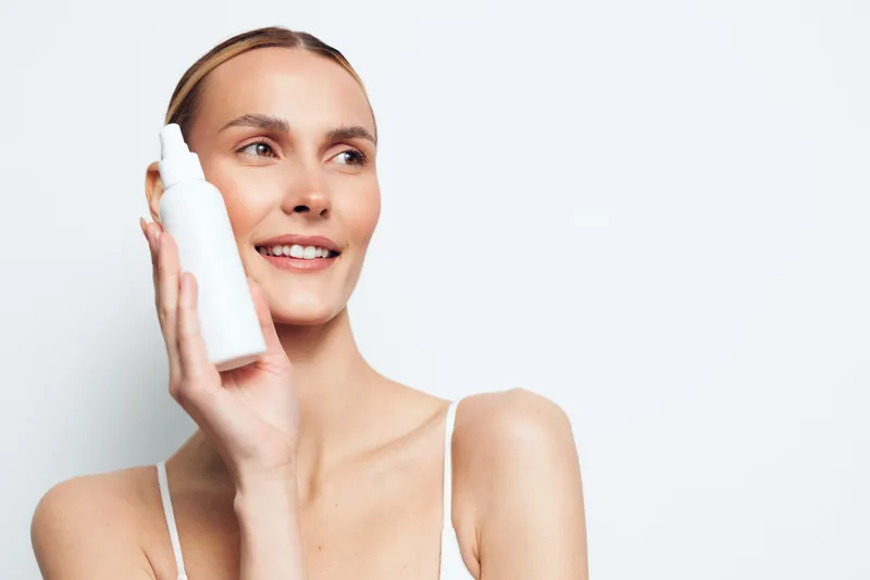 Smiling woman holding a bottle of moisturizer, close up on radiant skin, soft lighting, minimal makeup, clean background, fresh and confident skincare moment