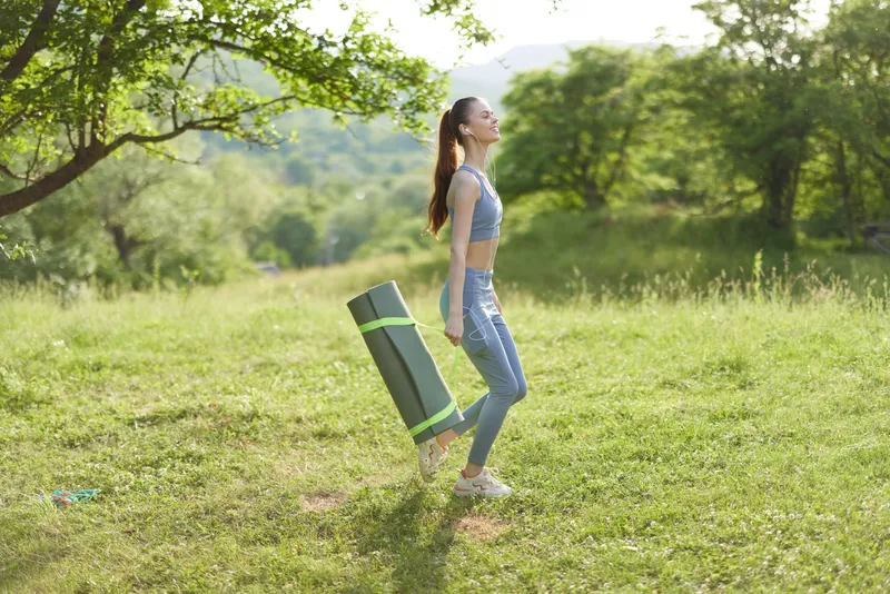 Young woman practicing yoga outdoors with mat in hand, enjoying nature, casual sportswear, vibrant green background, healthy lifestyle concept
