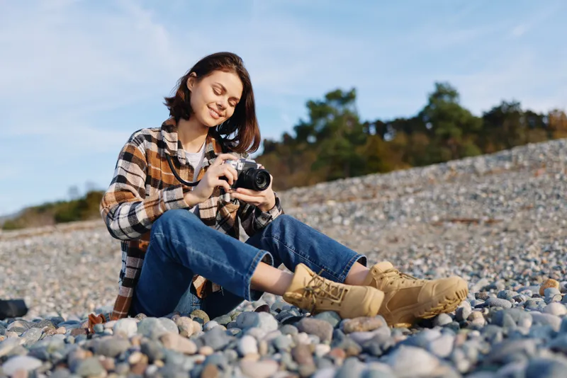 Woman photographer on a pebble beach, wearing a plaid shirt and boots, focused on her camera while capturing the landscape and textured shoreline around her