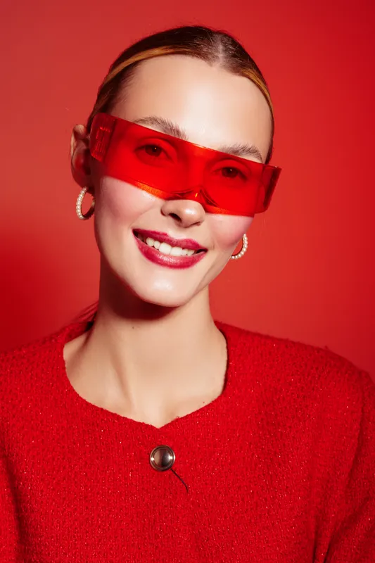 Smiling woman with red sunglasses and red sweater poses against red background. Fashion portrait with bright colors, modern style, and confident expression in studio lighting.