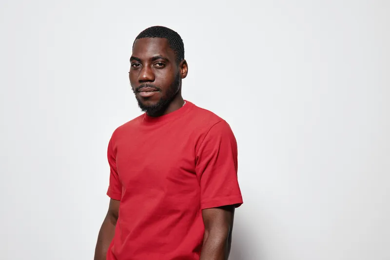 African American man with dark skin wearing red t shirt standing against white background looking at camera with confident expression and relaxed posture in studio.