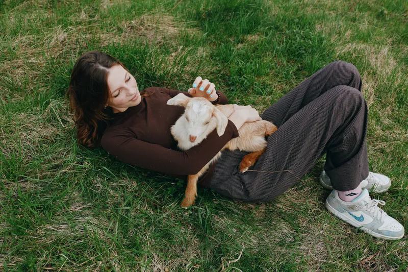 Relaxed woman lying on grass with a playful goat kid, sharing gentle affection in an outdoor lifestyle moment, casual outfit, calm countryside vibe, soft light and candid interaction