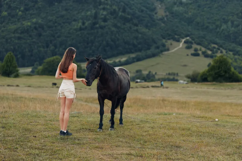 A young woman stands beside a horse in an open meadow with rolling hills in the background, calm and curious, sharing a quiet moment in nature.