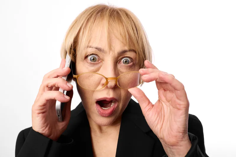 Elderly woman surprised closeup portrait adjusting glasses while holding a phone in studio, white background, astonished expression, comedic mood, shock gesture