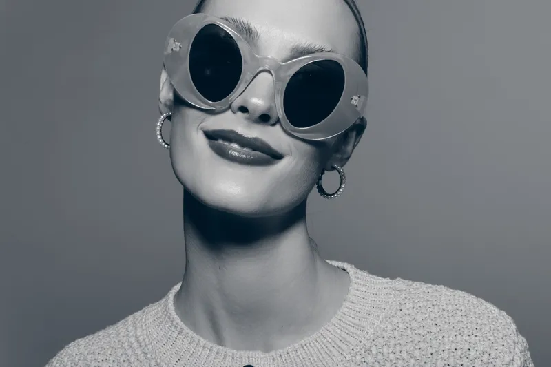 Young white woman with pale skin wearing large round sunglasses and hoop earrings smiles confidently in studio portrait. Concept of fashion, style, and modern accessories.