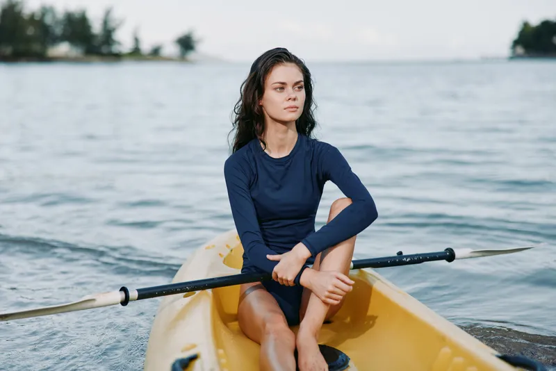 A young woman sits in a yellow kayak on calm water, grasping a double oar, wearing a navy long sleeve shirt. The image conveys focus, balance, and quiet preparation for movement.