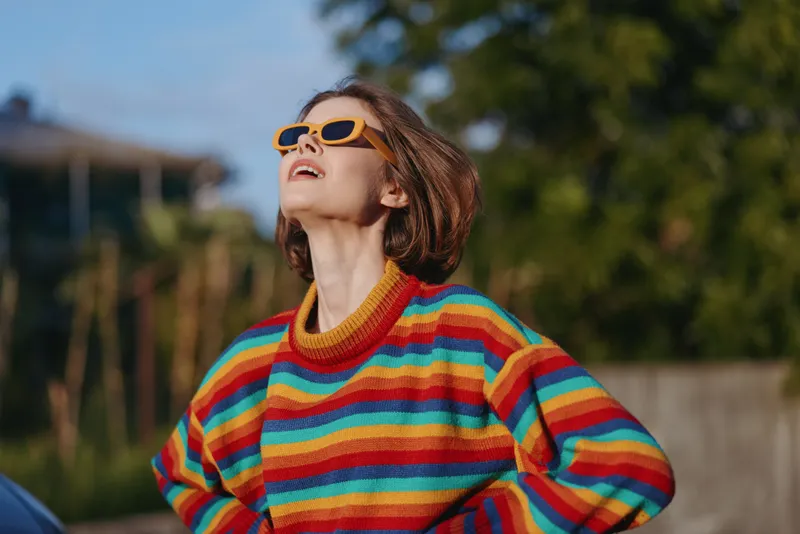 Woman in a striped sweater and sunglasses looking up outdoors, short hair smiling confidently with hands on hips in sunlit suburban greenery, rainbow fashion casual lifestyle portrait.