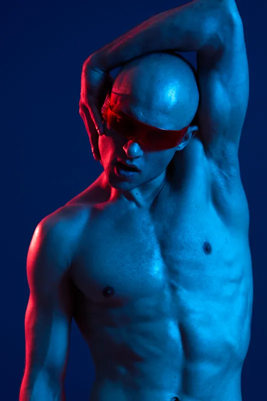 A striking man with a bald head poses dramatically, showcasing his sculpted physique against a vibrant blue backdrop. The image features bold lighting in shades of red and blue.
