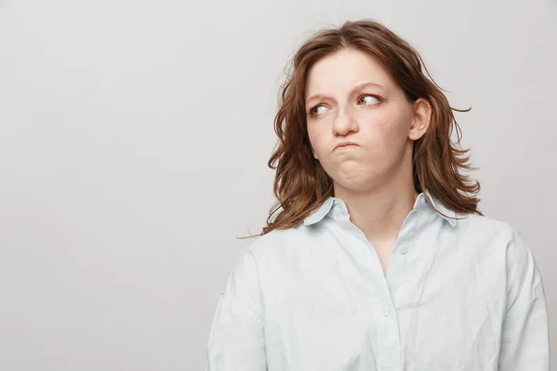 Young woman in white shirt looks sideways with skeptical expression on light background. Concept of doubt, suspicion, and uncertain emotions in casual portrait.