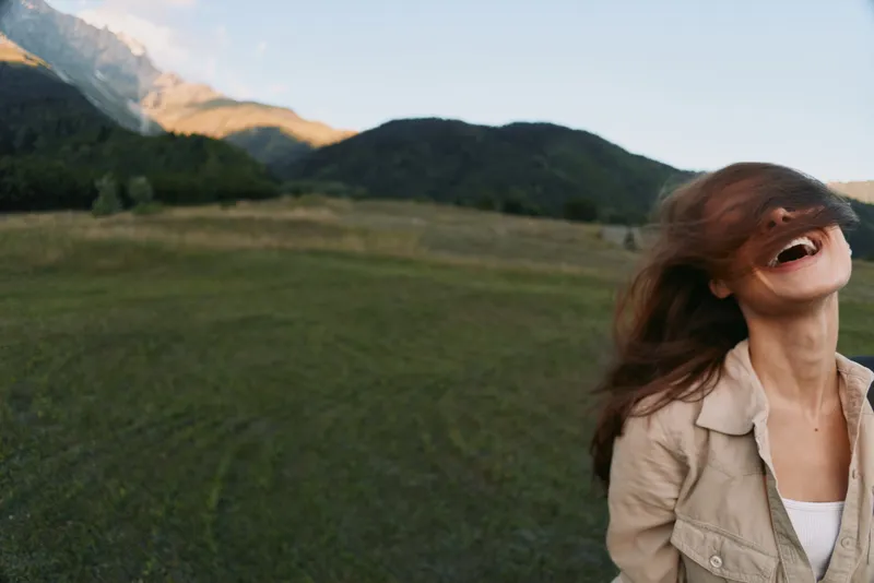 Happy woman with long hair laughing outdoors in a green field with mountains in the background. Natural light highlights a joyful lifestyle moment in nature.