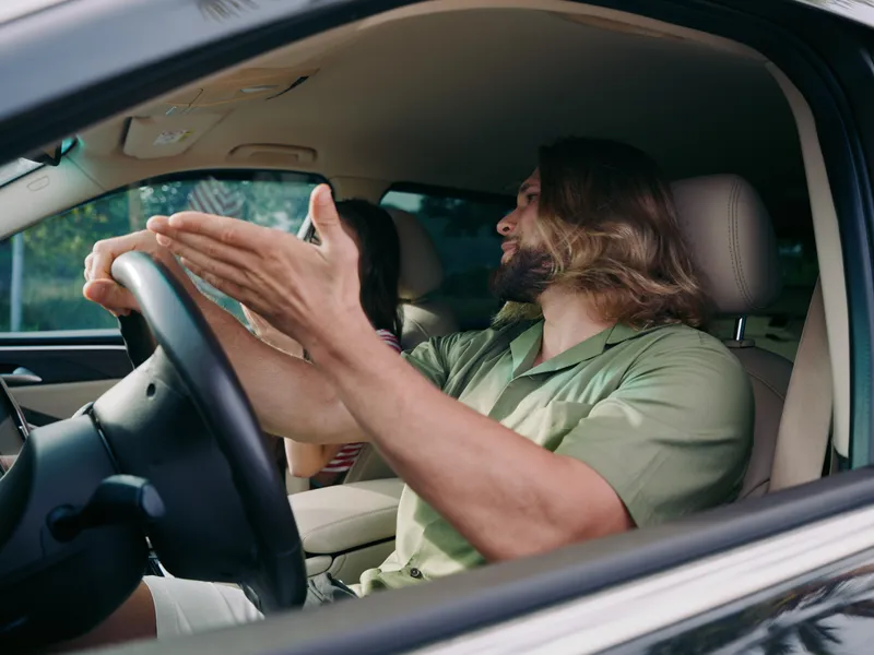 Close up of a bearded man driving a car, hands gripping the steering wheel, relaxed posture, casual shirt, illuminated interior, conveying focus, control, and motion.