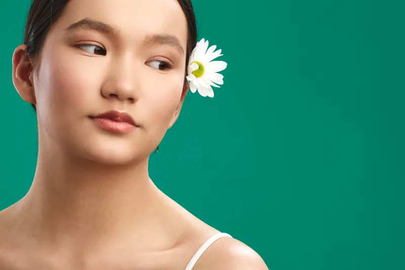 Asian woman with a calm expression gazes to the side, adorned with a white flower in her hair against a vibrant green backdrop, exuding graceful beauty and poise.