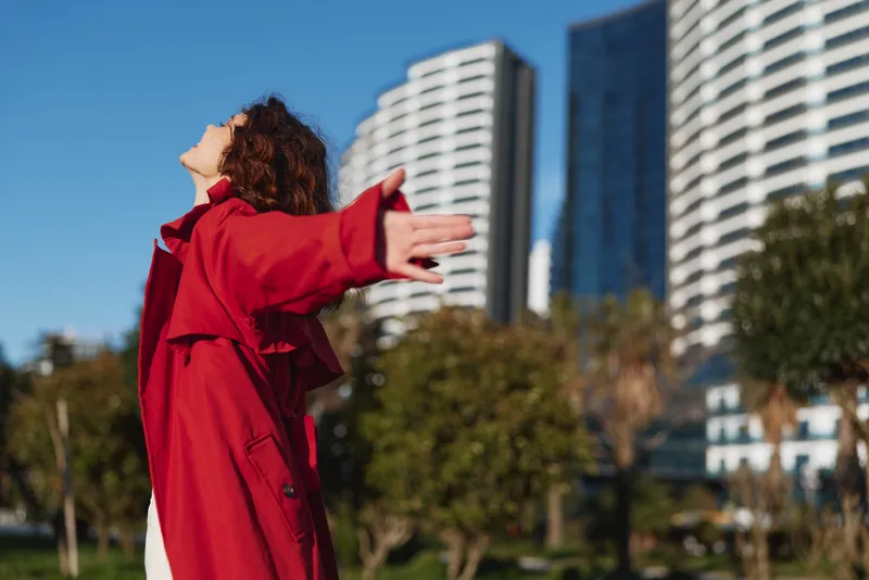 Young woman with curly hair wearing a red coat stands outdoors with arms outstretched enjoying a sunny day in an urban park setting.