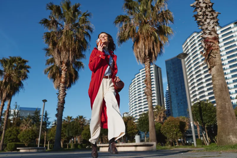 Young woman in a red coat walks confidently along a city street lined with palm trees on a sunny day, enjoying a moment of freedom and style.